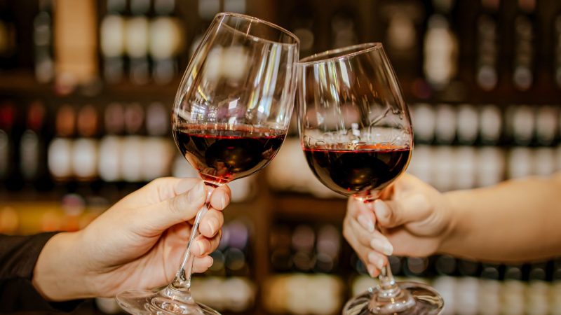 Brindis con dos copas de vino tinto en un restaurante, con estantería de botellas al fondo, ideal para una cena de San Valentín en Valencia.